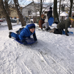 Chłopiec ubrany w niebieskie ubranie i czapkę leży na sankach w tle dzieci. Zjeżdżają w parku po górce pokrytej śniegiem. Kliknięcie na zdjęcie spowoduje jego powiększenie do rozmiaru oryginalnego.