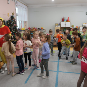 Przedszkolaki stoją w rzędzie przed starszymi koleżankami. Dziewczynki trzymają w ręce kartki i czytają im zagadki. Kliknięcie na zdjęcie spowoduje jego powiększenie do rozmiaru oryginalnego.