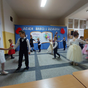 Przedszkolaki tańczą trzymając się za ręce. W tle dekoracja z okazji Dnia Babci i Dziadka. Kliknięcie na zdjęcie spowoduje jego powiększenie do rozmiaru orygianlnego.