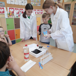 Uczennice z kółka chemicznego pokazują dzieciom ze świetlicy eksperyment z sodą. Dziewczynka trzyma torebkę w ręku obok niej chłopiec, na stoliku sprzęt chemiczny. Kliknięcie na zdjęcie spowoduje jego powiększenie do rozmiaru oryginalnego.