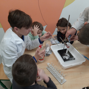 Uczeń ubrany w fartuch laboratoryjny pokazuje dzieciom przebywającym na świetlicy eksperymenty. Na stoliku sprzęt chemiczny potrzebny do doświadczenia. Kliknięcie na zdjęcie spowoduje jego powiększenie go do rozmiaru oryginalnego.