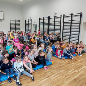 Przedszkolaki i dzieci z kl. 1-3 uczestniczą w spotkaniu z policjantem. Dzieci siedzą zgromadzone na sali gimnastycznej. Kliknięcie na zdjęcie spowoduje jego powiększenie do rozmiaru oryginalnego.