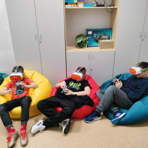Uczniowie siedzą na kolorowych pufach, za nimi szafki. Młodzież ogląda bryły matematyczne w okularach VR. Kliknięcie na zdjęcie spowoduje jego powiększenie do rozmiaru oryginalnego.