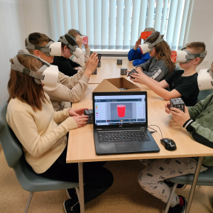 Uczniowie kl. 6a oglądają w okularach VR bryły matematyczne. Siedzą przy stolikach na głowach mają okulary VR. Na ekranie laptopa model walca. Kliknięcie na zdjęcie spowoduje jego powiększenie do rozmiaru oryginalnego.