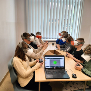 Uczniowie siedzą w bibliotece szkolnej. Na głowie mają okulary VR w rękach trzymają kody QR. Obracając kostką oglądają modele 3D brył matematycznych. Na stoliku laptop z modelem kuli 3D. Kliknięcie na zdjęcie spowoduje jego powiększenie do rozmiaru oryginalnego.