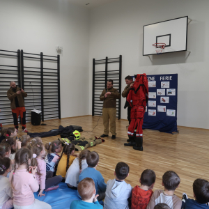 Jeden ze strażaków pokazuje uczniom czerwony kombinezon strażacki i omawia do czego służy. Kliknięcie na zdjęcie spowoduje jego powiększenie do rozmiaru oryginalnego.
