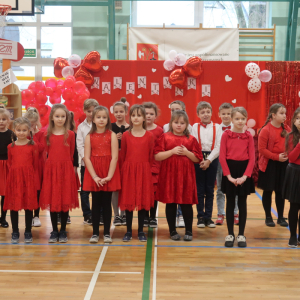 Uczniowie występują na scenie z okazji Walentynek. Dzieci stoją obok siebie ubrane w czerwone stroje w tle czerwona dekoracja z sercami. Kliknięcie na zdjęcie spowoduje jego powiększenie do rozmiaru oryginalnego.