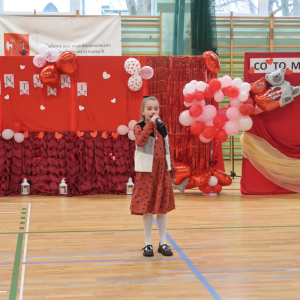 Kamila Górska z kl. 3 b śpiewa na hali gimnastycznej. Dziewczynka stoi na środku, w tle tematyczna walentynkowa dekoracja z balonami. Kliknięcie na zdjęcie spowoduje jego powiększenie do rozmiaru oryginalnego.