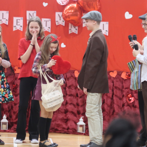 Uczniowie przebrani stoją na scenie. Dziewczynka trzyma siatkę i serce. Towarzyszą jej inni uczniowie. Kliknięcie na zdjęcie spowoduje jego powiększenie do rozmiaru oryginalnego.