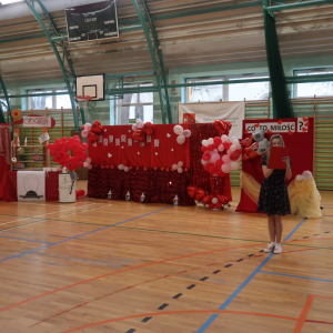 Uczennica kl. 5 a czyta tekst dotyczący Walentynek. Dziewczynka stoi na hali gimnastycznej. W tle dekoracja na czerwonych materiałach z balonami. Kliknięcie na zdjęcie spowoduje jego powiększenie do rozmiaru oryginalengo.