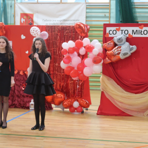 Uczennice stoją na środku hali gimnastycznej. Dziewczynki ubrane w czarne sukienki recytują "Hymn do miłości" św. Pawła. W tle czerwona walentynkowa dekoracja. Kliknięcie na zdjęcie spowoduje jego powiększenie do rozmiaru oryginalnego.