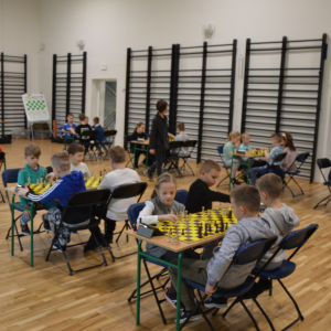Uczestnicy szkolnego turnieju szachowego siedzą na krzesełkach.Dzieci grają w szachy. Kliknięcie na zdjęcie spowoduje jego powiększenie do rozmiaru oryginalnego.