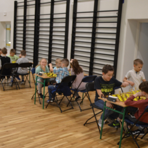 Dzieci z kl. 1-3 siedzą przy stolikach na sali gimnastycznej. Pomiędzy nimi szachownica. Grają w trunieju szachowym. Kliknięcie na zdjęcie spowoduje jego powiększenie do rozmiaru oryginalnego.