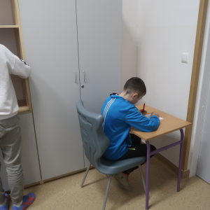 Uczeń zaznacza swoje głosy na karcie wyborczej. Chłopiec siedzi na krześle przy stoliku w bibliotece szkolnej.  Kliknięcie na zdjęcie spowoduje jego powiększenie do rozmiaru oryginalnego.