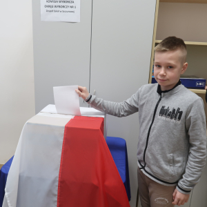 Uczeń klasy 6 a stoi z ręką wyciągniętą nad urną wyborczą znajdującą się w bibliotece szkolnej. Chłopiec wrzuca swoją kartę wyborczą.  Kliknięcie na zdjęcie spowoduje jego powiększenie do rozmiaru oryginalnego.