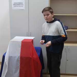 Uczeń kl. 6 b stoi obok urny wyborczej przykrytej biało-czerwoną flagą. Chłopiec trzyma w ręce kartę wyborczą zgiętą na pół. Kliknięcie na zdjęcie spowoduje jego powiększenie do rozmiaru oryginalnego.