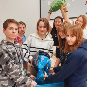 Szkolni wolontariusz trzymają plastikowe worki z nakrętkami. Uczniowie i uczennice stoją w grupie. Kliknięcie na zdjęcie spowoduje jego powiększenie do rozmiaru oryginalnego.