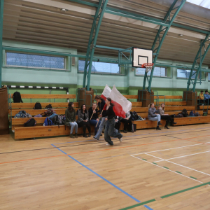 Uczennice biegają z flagami Polski. Uczestniczą na hali gimnastycznej w biegu upamiętniającym Żołnierzy Wyklętych. Na trybunach sportowych siedzą nauczyciele i pozostali uczniowie. Kliknięcie na zdjęcie spowoduje jego powiększenie do rozmiaru oryginalnego.