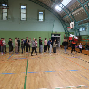 Uczniowie uczestniczą w biegu międzyklasowym. Biegają wokoło hali gimnastycznej z flagami Polski w rękach. Pozostali uczniowie stoją w parach i czekają na swoją kolej. Kliknięcie na zdjęcie spowoduje jego powiększenie do rozmiaru oryginalnego.