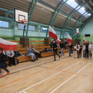Uczniowie biegają z flagami Polski w rękach na hali gimnastycznej Zespołu Szkół w Szczurowej. Młodzież upamiętnia Żołnierzy Wyklętych. Towarzyszom im nauczyciele siedzący na trybunach. Kliknięcie na zdjęcie spowoduje jego powiększenie do rozmiaru oryginalnego.