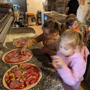 Dziewczynki układają szynkę oraz inne składniki na pizzy. Uczestniczą w warsztatach kulinarnych w pizzerii "Giona". Kliknięcie na zdjęcie spowoduje jego powiększenie do rozmiaru oryginalnego.