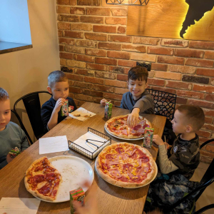 Czterech chłopców siedzi przy stole na krzesełkach. Przed nimi na okrągłych talerzach pizza. Przedszkolaki jedzą pizzę. Kliknięcie na zdjęcie spowoduje jego powiększenie do rozmiaru oryginalnego.