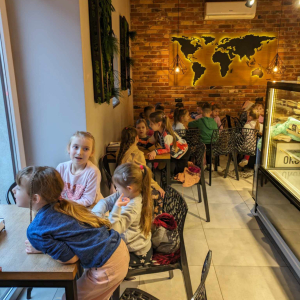 Dzieci siedzą w pizzerii "Giona" przy drewnianych stolikach na czarnych krzesełkach. W tle kontury kontynentów na ceglanej ścianie. Kliknięcie na zdjęcie spowoduje jego powiększenie do rozmiaru oryginalnego.