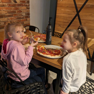 Dwie dziewczynki siedzą w pizzerii "Giona" przy drewnianych stolikach. Jedzą przygotowaną przez siebie pizzę. Na stole na talerzu upieczona pizza. Kliknięcie na zdjęcie spowoduje jego powiększenie do rozmiaru oryginalnego.
