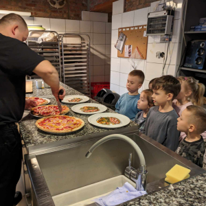Przedszkolaki stoją w kuchni pizzerii "Giona" przy blacie kuchennym. Obserwują jak pan Konrad - kucharz kroi upieczone pizzę. Kliknięcie na zdjęcie spowoduje jego powiększenie do rozmiaru oryginalnego.