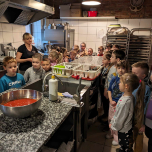 Dzieci stoją wokół blatu kuchennego w pizzerii "Giona". Towarzyszy im pracownica pizzerii. Na blacie misa z sosem do pizzy oraz inne sprzęty kuchenne. Kliknięcie na zdjęcie spowoduje jego powiększenie do rozmiaru oryginalnego.