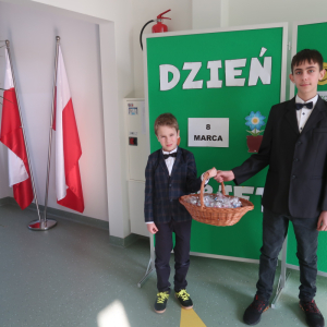 Przedstawiciele Samorządu Uczniowskiego stoją na korytarzu. W rękach trzymają kosz ze słodkimi upominkami. W tle dekoracja i flagi Polski. Kliknięcie na zdjęcie spowoduje jego powiększenie do rozmiaru oryginalnego.