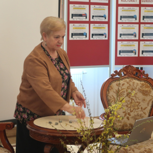 Pani Irena Góra prezentuje na stole zabawę z pięcioma kamieniami. Obok stolika zabytkowe krzesło, w tle stelaże z gminnymi informacjami. Kliknięcie na zdjęcie spowoduje jego powiększenie do rozmiaru oryginalnego.
