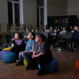 Wychowankowie świetlicy siedzą na pufach i krzesełkach w Gminnym Centrum Kultury Czytelnictwa i Sportu. Dzieci oglądają film o Minionkach wraz z opiekunami. Kliknięcie na zdjęcie spowoduje jego powiększenie do rozmiaru oryginalnego.