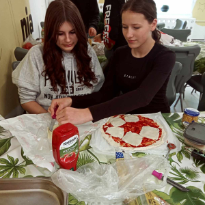 Uczennice kl. 7 a na szkolnej stołówce przygotowują swoją pizzę. Dziewczynki siedzą przy stoliku i nakładają składniki na ciasto. Kliknięcie na zdjęcie spowoduje jego powiększenie do rozmiaru oryginalnego.