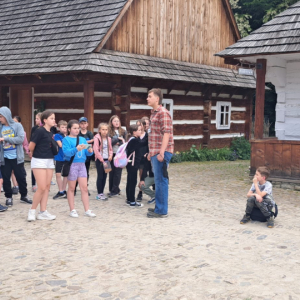 Uczniowie zwiedzają Muzeum Budownictwa Ludowego w Sanoku wraz z opiekunami i przewodnikiem. Stoją obok muzealnych obiektów.  Kliknięcie na zdjęcie spowoduje jego powiększenie do rozmiaru oryginalnego.