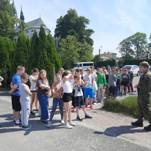 Uczniowie zebrani przed parkingiem szkolnym. Uczestniczą w spotkaniu z żołnierzem.  Kliknięcie na zdjęcie spowoduje jego powiększenie do rozmiaru oryginalnego.