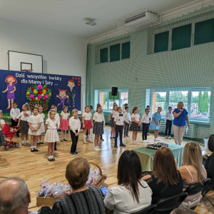 Uczniowie kl. 2 ubrani w kolorowe stroje prezentują przedstawienie dla swoich mam i ojców. Obok dzieci zebranych na sali gimnastycznej stoi pani Iwona Czesaki i przemawia do mikrofonu. Kliknięcie na zdjęcie spowoduje jego powiększenie do rozmiaru oryginalnego.