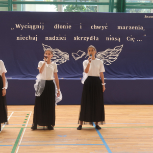 Absolwentki kl. 8 w biało-czarnych strojach wykonują piosenkę. W tle dekoracja z białym napisem, skrzydłami i czapką absolwenta.  Kliknięcie na zdjęcie spowoduje jego powiększenie do rozmiaru oryginalnego.