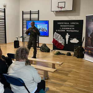 Żołnierz przedstawia uczniom specyfikę pracy wojskowej. W tle tematyczna dekoracja i ekran multimedialny. Kliknięcie na zdjęcie spowoduje jego powiększenie do rozmiaru oryginalnego.