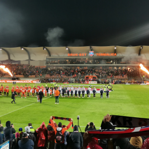 Piłkarze Bruk-Bet Termalica i Wisły Kraków na murawie stadionu. Wokół trybuny z kibicami wśród, których znajdują się wychowankowie naszej szkoły razem z rodzicami. Kliknięcie na zdjęcie spowoduje jego powiększenie do rozmiaru oryginalnego.