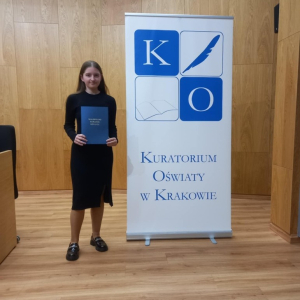 Laureatka Małopolskiego Konkursu Języka Polskiego - Dorota Budzioch z kl. 8 a  z nagrodą przy banerze z logotypem Małopolskiego Kuratorium Oświaty w Krakowie. Kliknięcie na zdjęcie spowoduje jego powiększenie do rozmiaru oryginalnego.