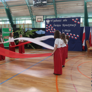 Dziewczynki z kl. 4 a trzymają kawałki materiału w kolorach białych i czerwonych. Wykonują taniec patriotyczny. Uczennica występuje na akademii z okazji Święta Konstytucji 3 Maja. Kliknięcie na zdjęcie spowoduje jego powiększenie do rozmiaru oryginalnego.