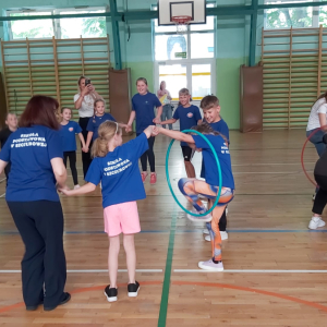 Dzieci i rodzice uczniów klas drugich oraz nauczyciele przekładają kółko hula-hop w czasie zabawy na hali gimnastycznej. Kliknięcie na zdjęcie spowoduje jego powiększenie do rozmiaru oryginalnego.