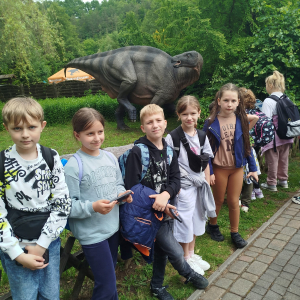 Grupka uczniów w parku dinozaurów. W tle model jednego z dinozaurów. Kliknięcie na zdjęcie spowoduje jego powiększenie do rozmiaru oryginalnego.