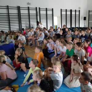 Dzieci wraz z nauczycielami słuchają prelekcji podróżnika na sali gimnastycznej.  Kliknięcie na zdjęcie spowoduje jego powiększenie do rozmiaru oryginalnego.