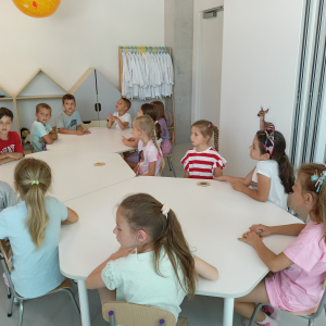 Dzieci siedzą przy stolikach w tle drewniane szafki w kształcie domków. Znajdują się w Centrum Cogiteon w Krakowie. Kliknięcie na zdjęcie spowoduje jego powiększenie do rozmiaru oryginalnego.