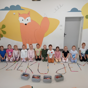 Dzieci ze świetlicy siedzą na podłodze, przed nimi napis ułożony z kredek przekazanych przez starszych uczniów. Kliknięcie na zdjęcie spowoduje jego powiększenie do rozmiaru oryginalnego.