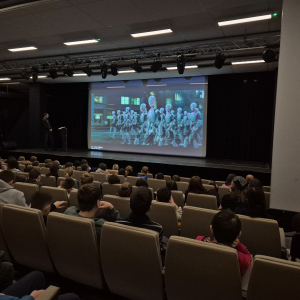 Młodzież w trakcie lekcji o AI, na ekranie roboty, na scenie osoba prowadząca zajęcia z uczniami.  Kliknięcie na zdjęcie spowoduje jego powiększenie do rozmiaru oryginalnego.