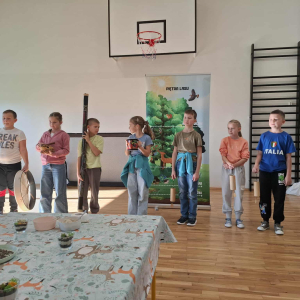 Uczniowie z klas 3 stoją na środku sali gimnastycznej z różnymi drewnianymi przedmiotami. W tle baner z lasem. Kliknięcie na zdjęcie spowoduje jego powiększenie do rozmiaru oryginalnego.