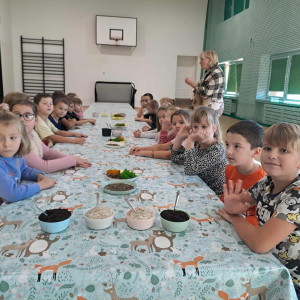 Klasy 1 w trakcie przygotowywania leśnych kompozycji. Uczniowie siedzą na krzesełkach przy stolikach. Kliknięcie na zdjęcie spowoduje jego powiększenie do rozmiaru oryginalnego.